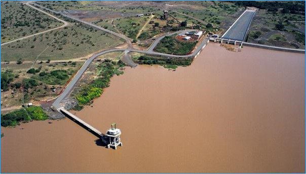 Aerial view of a large dam and intake tower connected to a reservoir with brownish water, featuring adjacent infrastructure and a long canal structure in a semi-arid landscape."